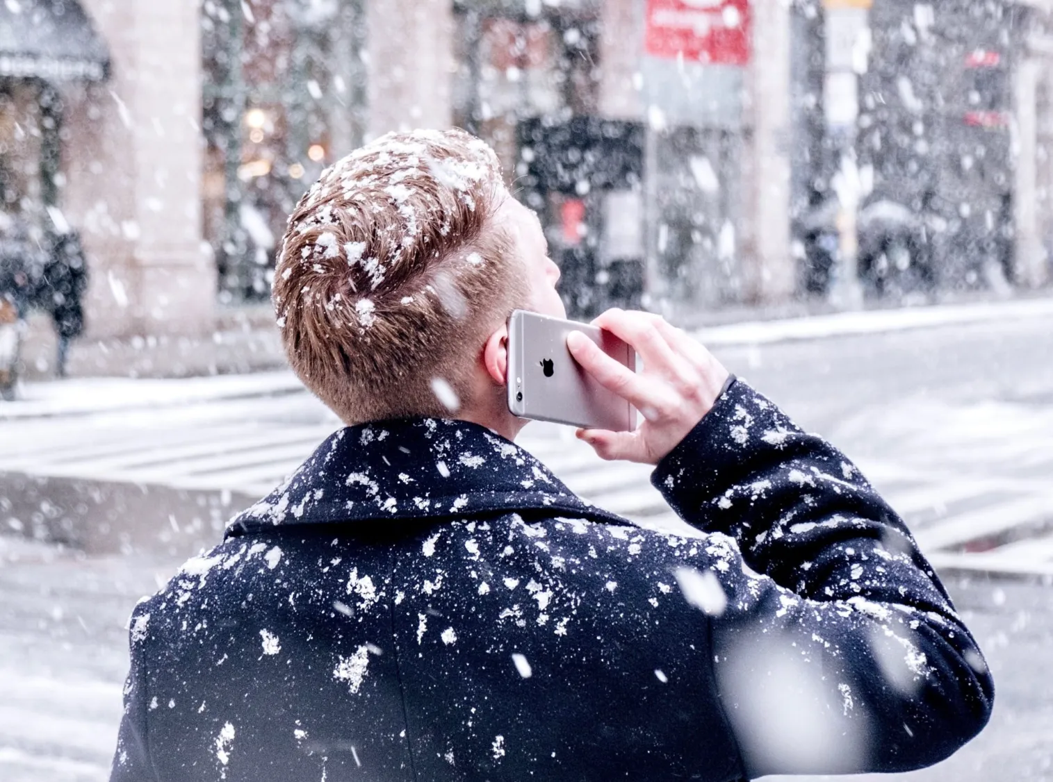 A blonde man with a short hair cut, on the phone outside during a snowy day.